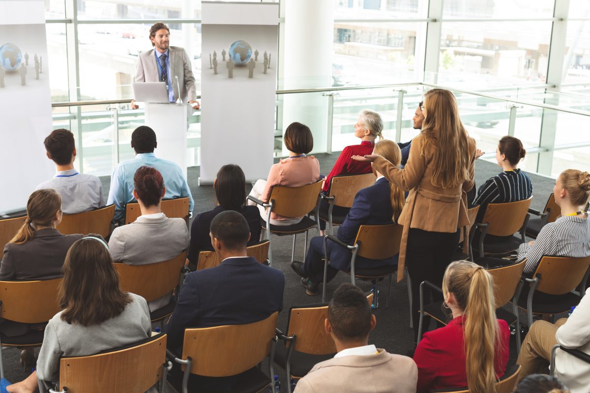 High angle view of Caucasian businesswoman interacting with Caucasian businessman speaking in front of business people sitting at business seminar in modern office building